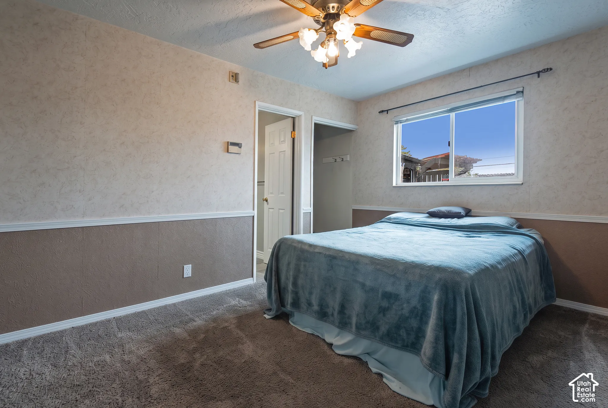 Bedroom featuring two-tone wall texturing with chair rail molding, wall-to-wall carpeting, sliding window with curtain rod, and a multi-bulb ceiling fan with wooden blades