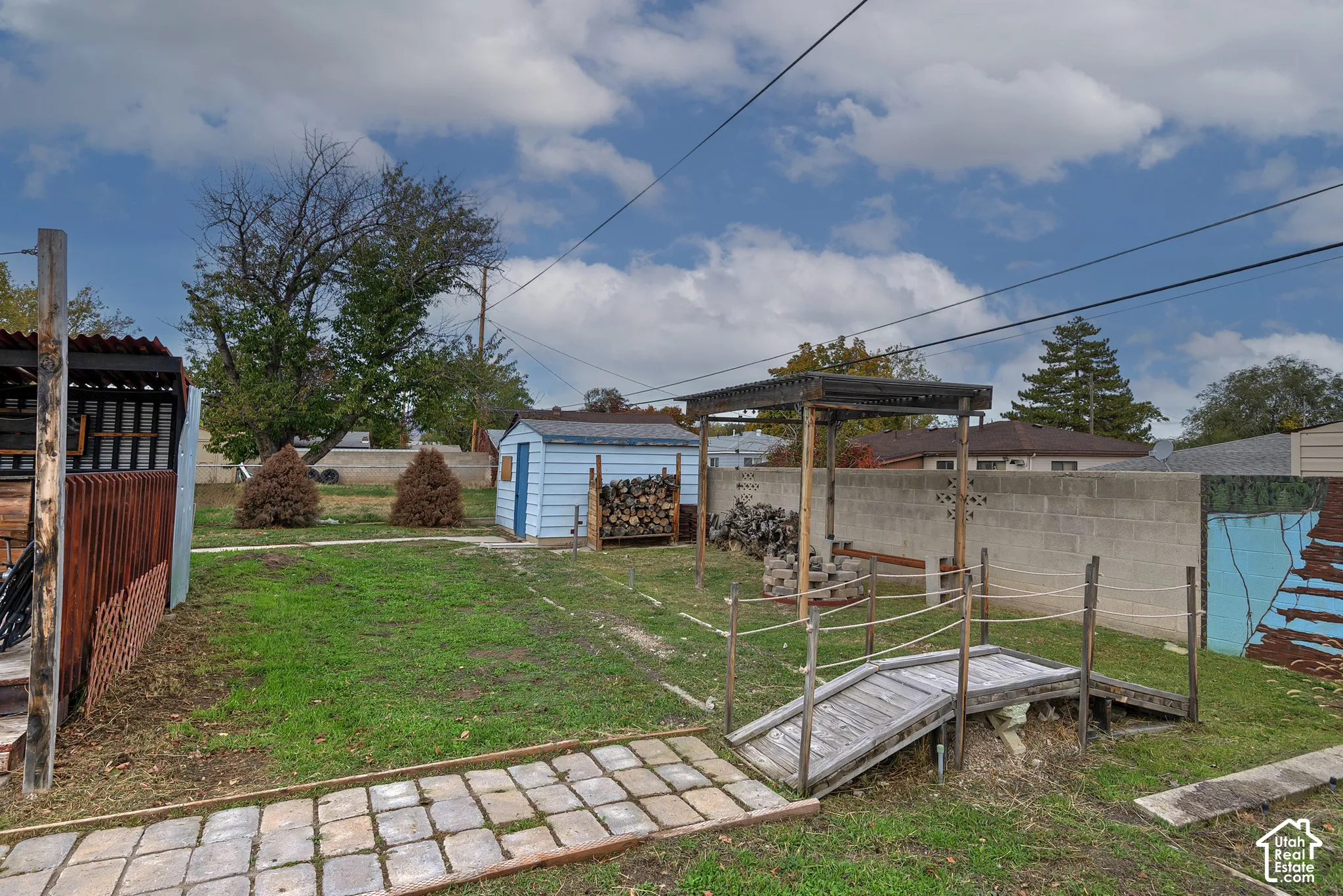 Rear exterior featuring a paved stone walkway, wooden footbridge, overhead pergola, detached storage structure, and concrete masonry perimeter wall
