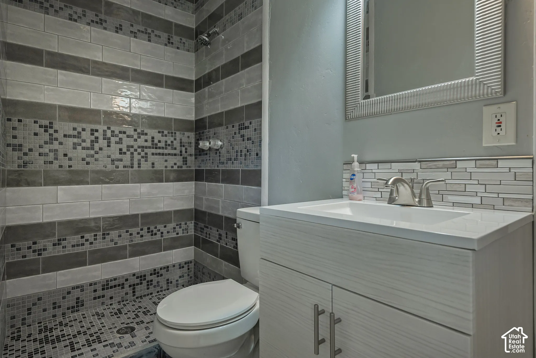 Walk-in shower with multi-tonal subway and mosaic tile surround, vanity cabinet featuring a neutral finish, integrated sink, and metallic hardware