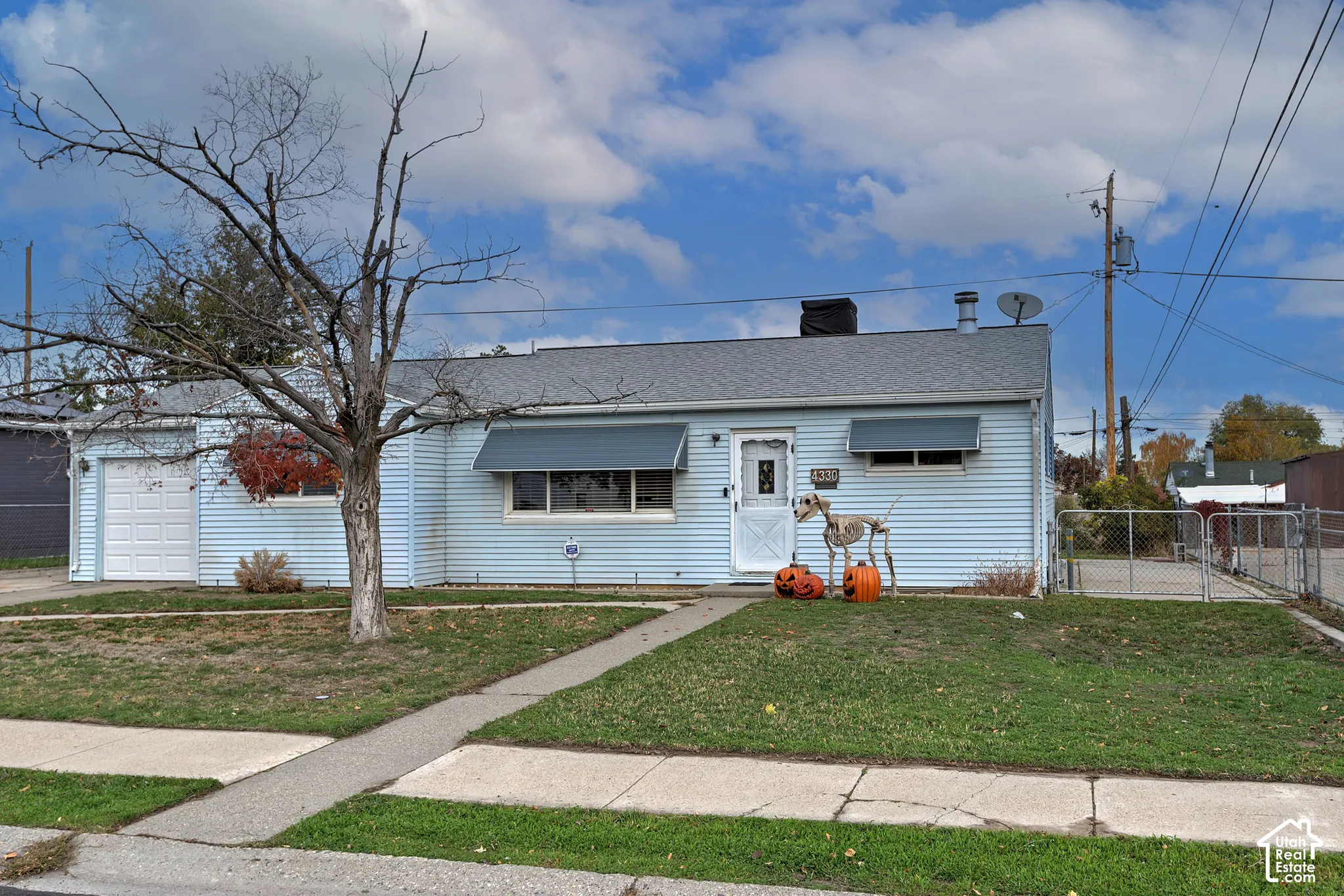 Single-story residence featuring light blue siding, an attached garage, metal window awnings, and a chain-link fenced side yard