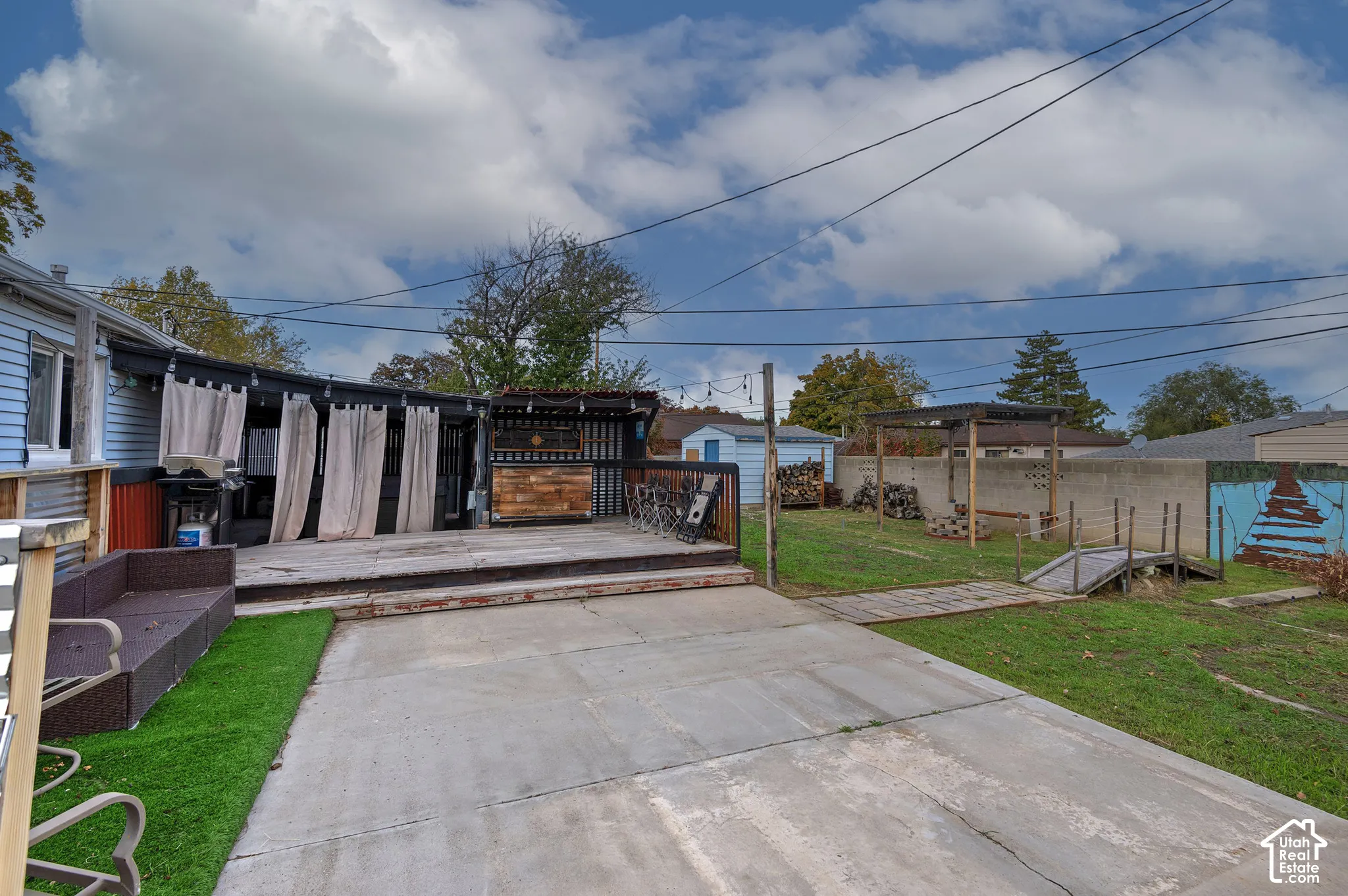 Rear exterior featuring a concrete patio, elevated wood-decking platform, integrated built-in bar, masonry perimeter wall, and turf-surfaced area