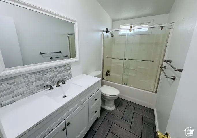 Bathroom featuring a white vanity with a rectangular basin, multi-toned stone tile backsplash, herringbone-patterned flooring, and a tub-shower combination enclosed by a sliding glass door