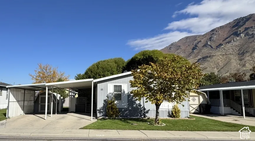 Exterior featuring light-toned vertical siding, a covered carport, and an attached storage shed, set against a prominent mountain backdrop