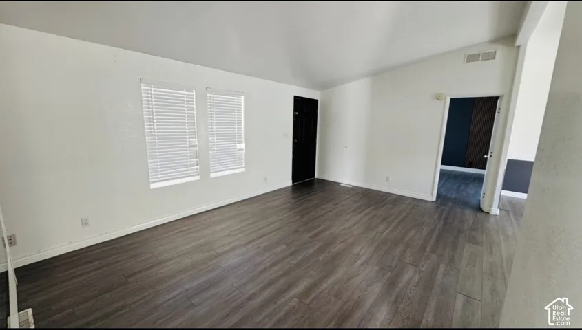 Vaulted ceilings, wood-finish flooring, and white walls throughout the space, featuring dual windows with horizontal blinds and a solid-panel door