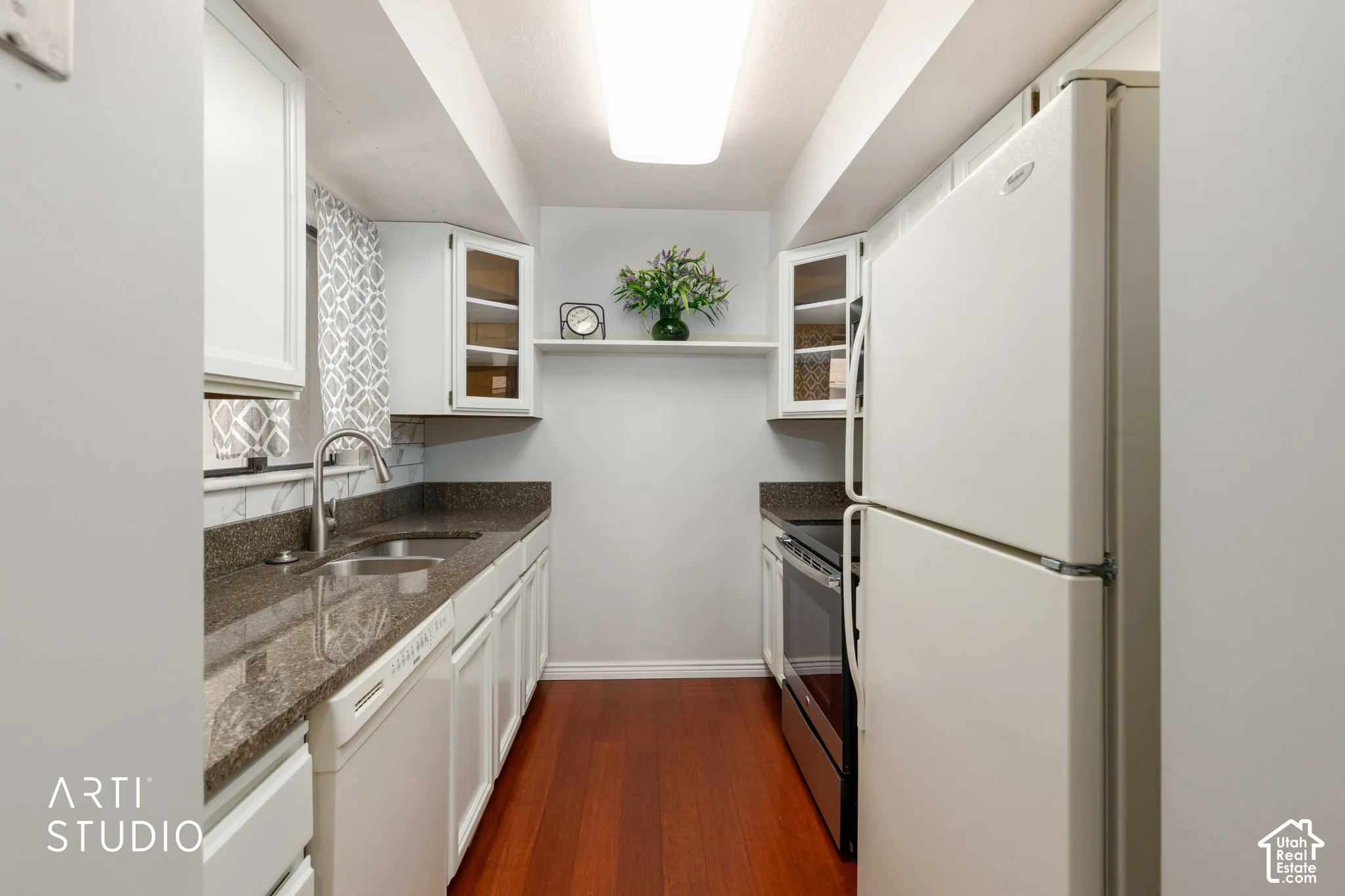 Galley kitchen featuring white cabinetry, stone-finish countertops, stainless steel dual-basin sink, integrated floating shelving, and wood-finish flooring