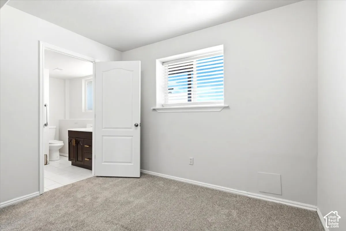 Room featuring neutral-toned carpeting, a window with horizontal blinds, and a paneled interior door opening to a bathroom equipped with a vanity and tile flooring