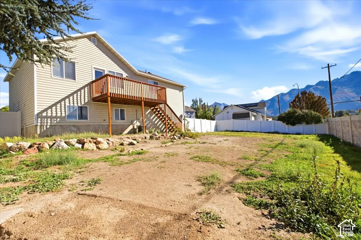 Exterior featuring light-toned horizontal siding, a raised wood deck with staircase, rock landscaping borders, and a perimeter vinyl fence set against a mountain backdrop