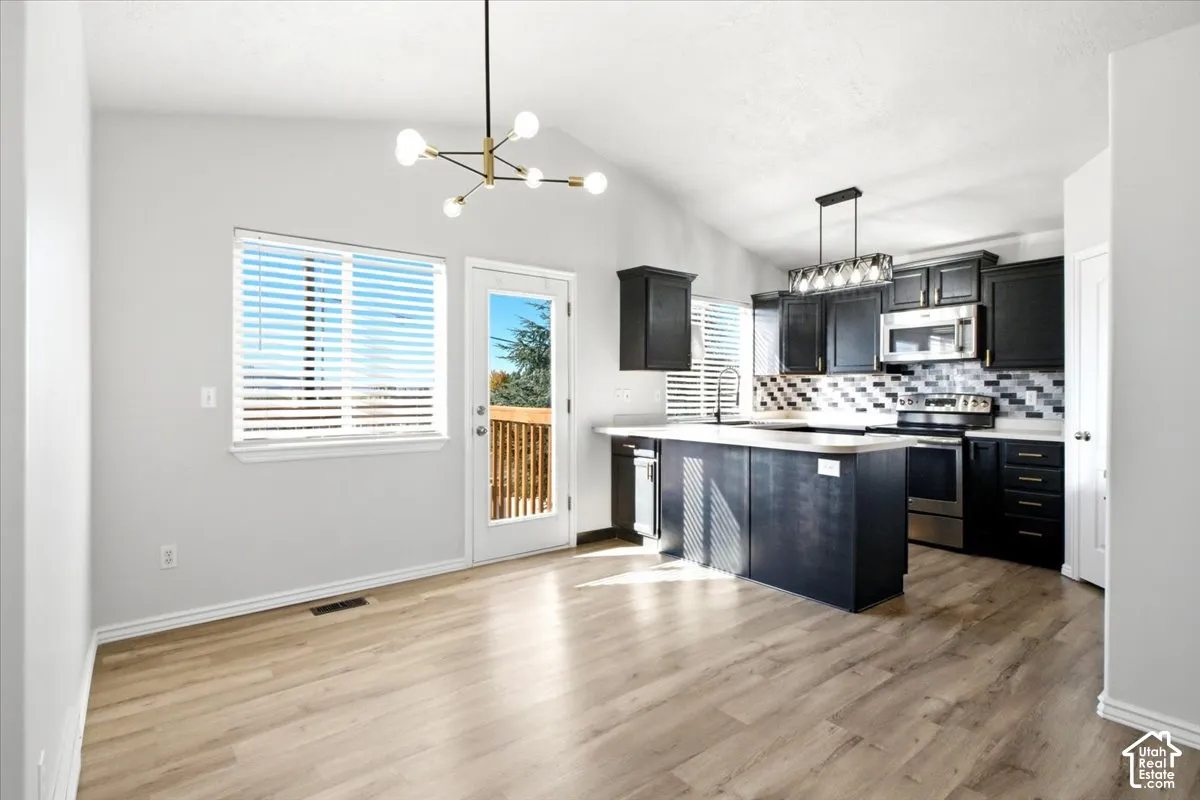 Open-concept interior featuring vaulted ceilings, wood-finish flooring, dark cabinetry, a multi-tone tile backsplash, and a modern geometric chandelier