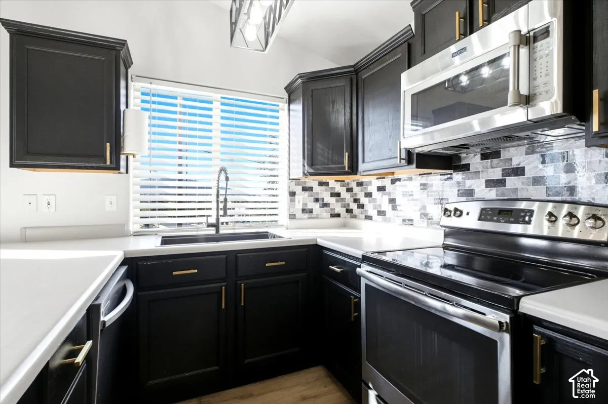 Kitchen featuring dark cabinetry with gold-tone hardware, stone-composite countertops, subway-style tile backsplash, stainless steel appliances, and wood-finish flooring
