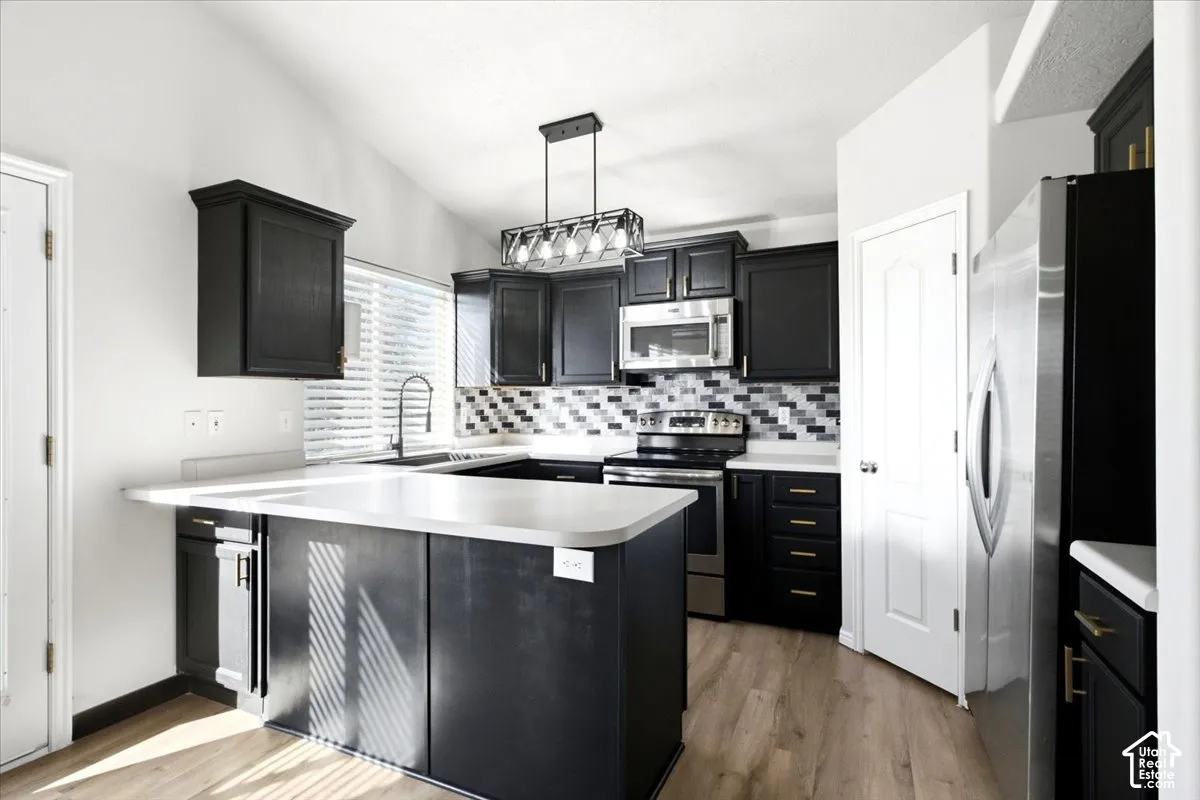 Kitchen featuring dark cabinetry, solid-surface countertops, multi-toned tile backsplash, stainless steel appliances, and a linear pendant light fixture