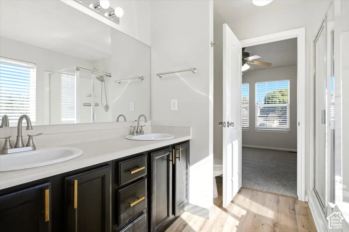 Dual-sink vanity with dark-finish cabinetry, gold-tone hardware, light-toned countertop, large wall-mounted mirror, and wood-finish flooring