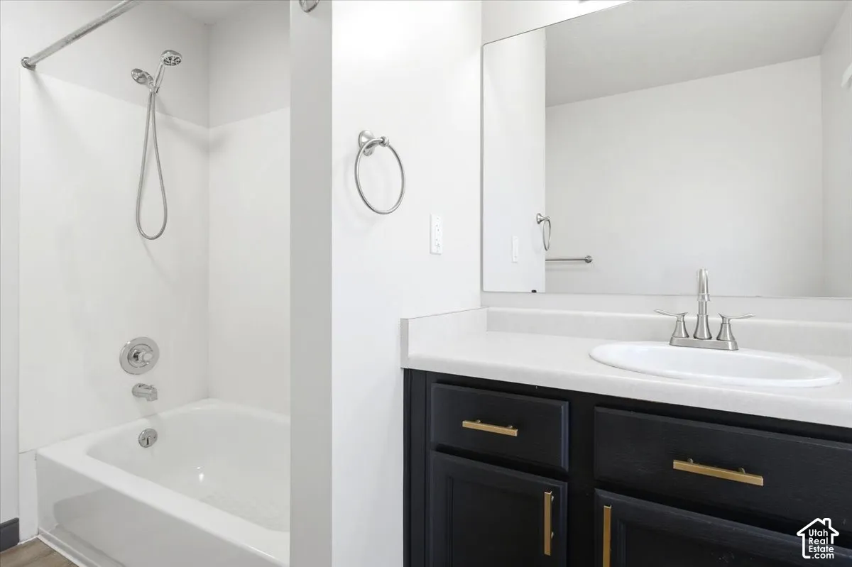 Bathroom featuring a dark wood-finish vanity, light solid-surface countertop with an integrated oval sink, matte gold hardware, wall-to-wall mirror, and a tub-shower combination with a hand-held sprayer