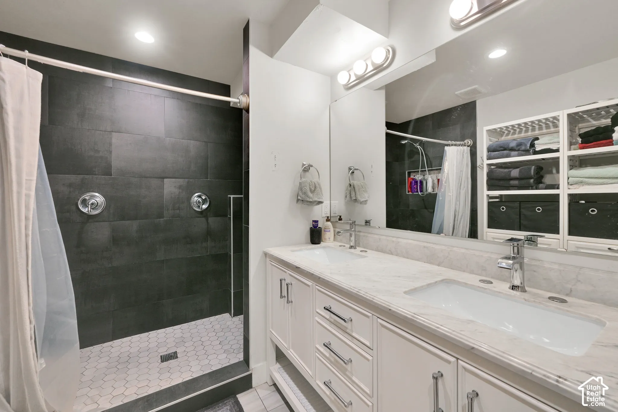 Double-vanity bathroom featuring large-format dark tile shower surrounds, white cabinetry with stone-finish countertops, undermount sinks, and full-width vanity mirrors