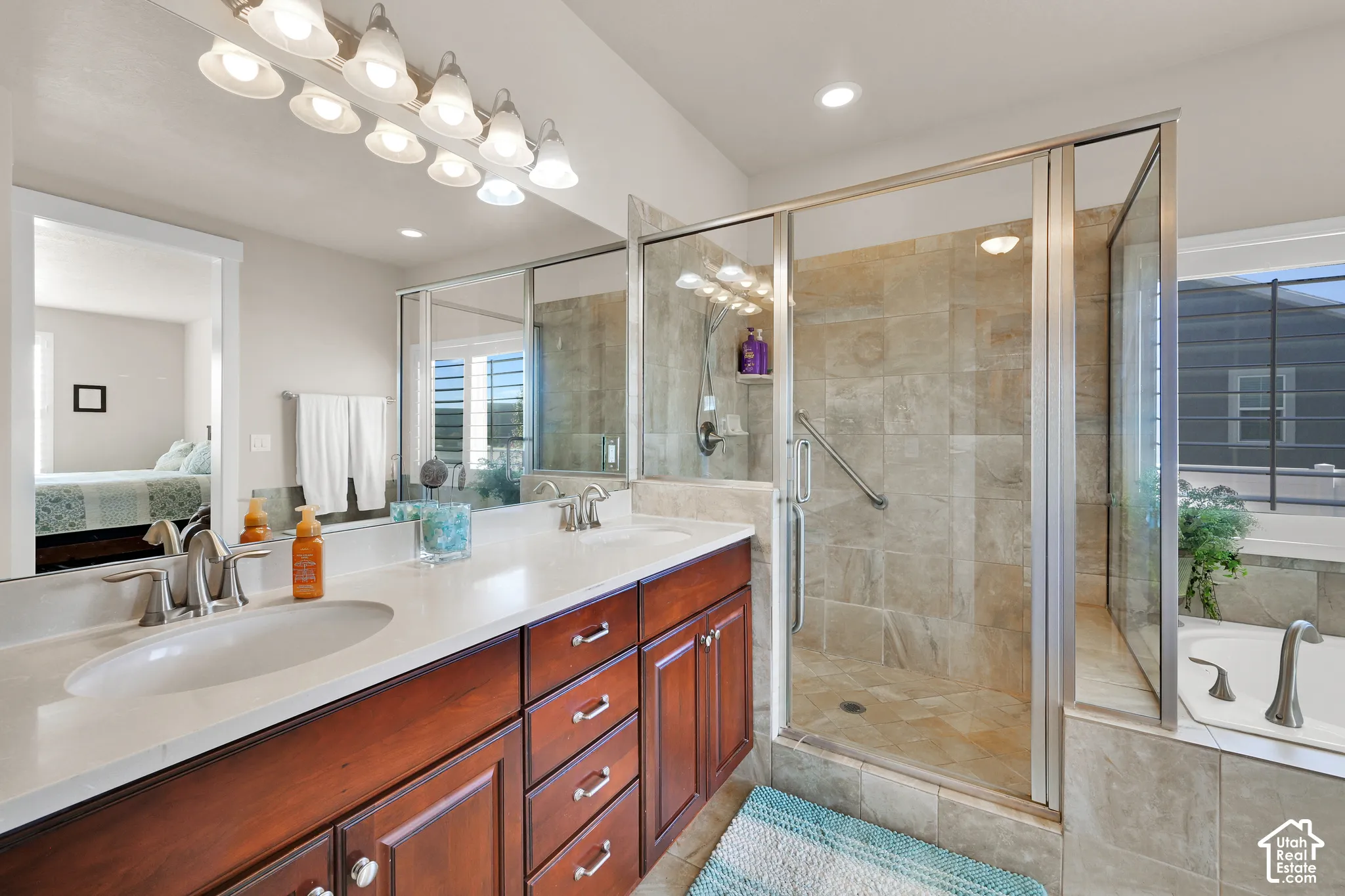 Dual-sink vanity with light-tone countertops and wood cabinetry, expansive wall-mounted vanity mirror, glass-enclosed shower with tile surround, and separate soaking tub