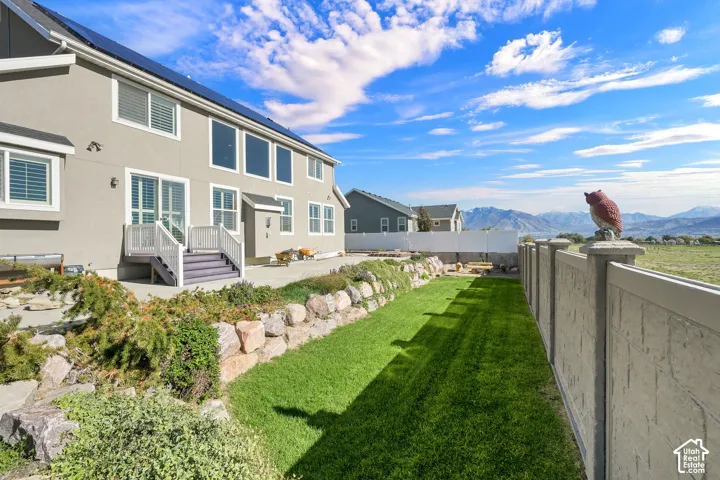 Rear exterior featuring a concrete patio with stairs, tiered rock retaining wall, manicured lawn, and perimeter fencing with mountain scenery
