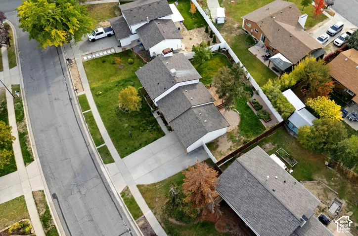 Aerial perspective of a property featuring a multi-pitched shingled roof, a concrete driveway, a paved circular patio, and mature trees within a fenced perimeter