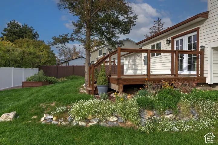 Raised wood deck featuring wire balusters, stone-lined garden beds, vinyl privacy fencing, and exterior wall-mounted lighting