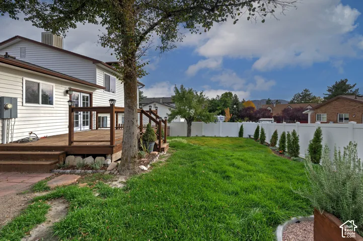 Rear exterior featuring a raised wood-finish deck with stairs, white vinyl privacy fencing, landscaped borders, and a lawn