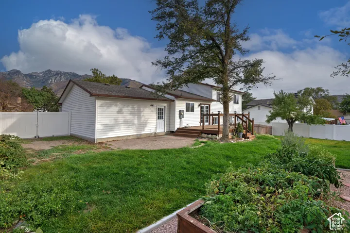 Rear exterior featuring white horizontal siding, raised wood deck, paver patio, white vinyl perimeter fencing, and mountain backdrop
