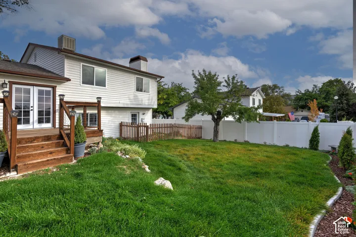 Backyard featuring a raised wooden deck with staircase, manicured lawn, stone landscaping borders, and a perimeter vinyl fence