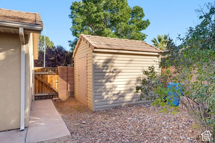 Exterior storage shed featuring a gabled tile roof and horizontal siding, positioned on a gravel-surfaced lot with concrete walkway and wood-panel fencing
