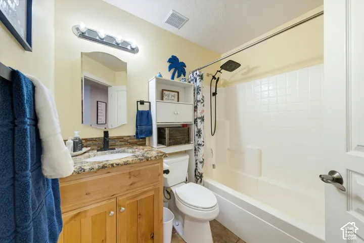 Bathroom featuring a wood cabinetry vanity, stone-patterned countertop, mosaic tile backsplash, oval-shaped mirror, and tub-shower combination with a square rainfall showerhead