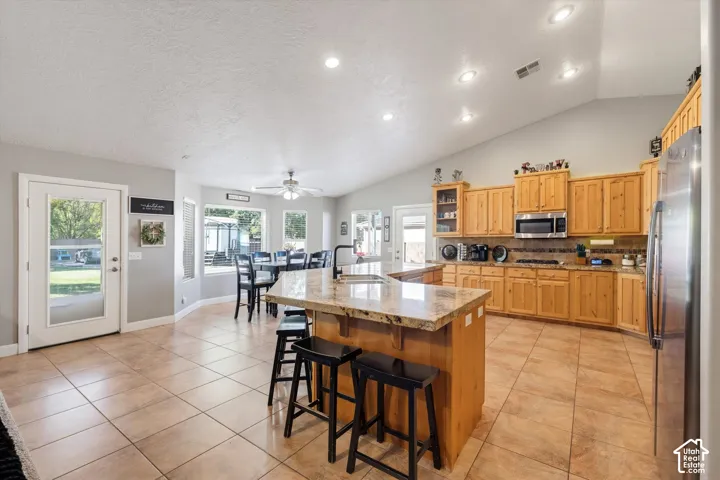 Open-concept kitchen featuring vaulted ceilings, recessed lighting, stone-surfaced island with integrated sink, natural wood cabinetry, and ceramic tile flooring