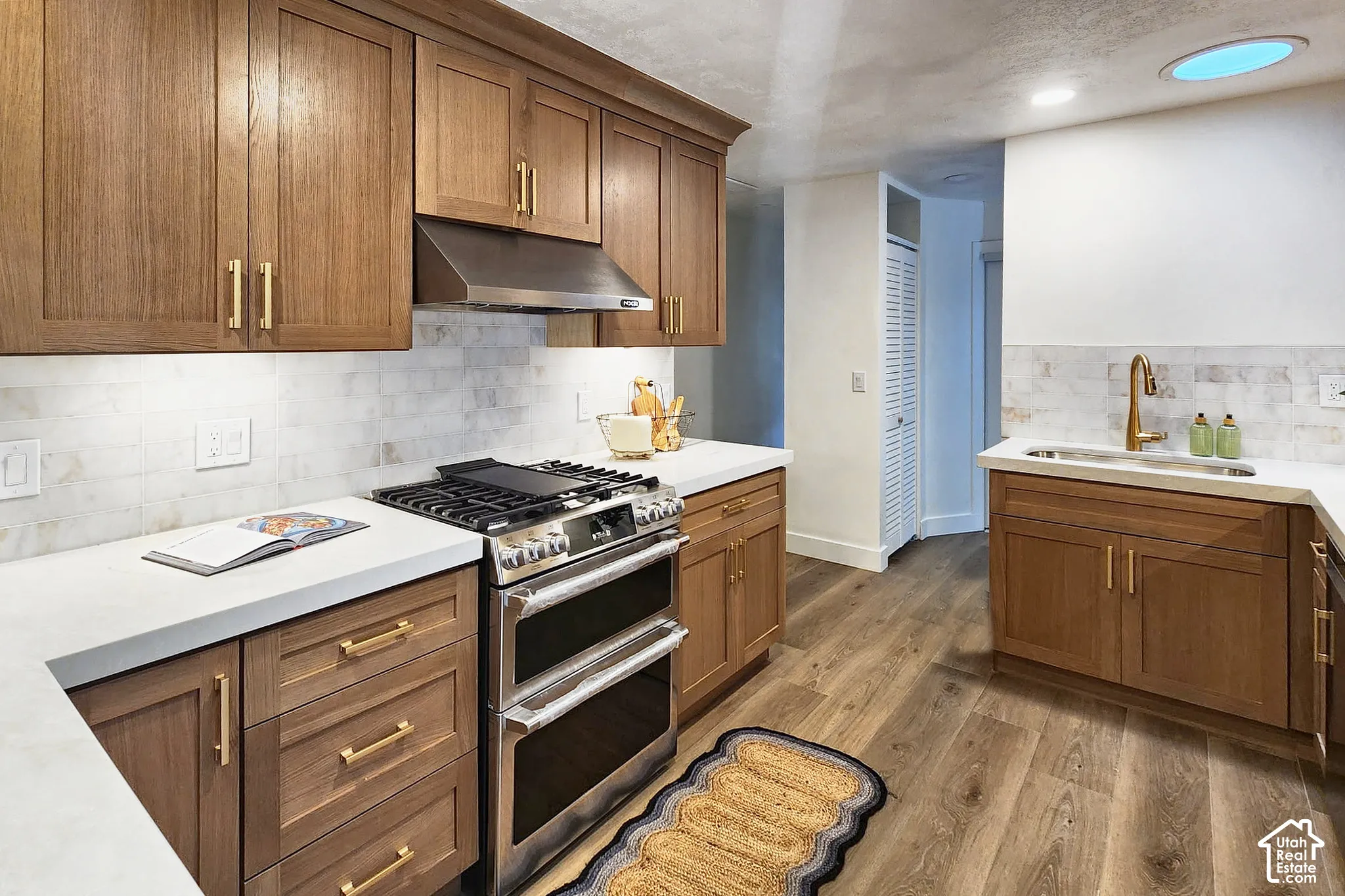Kitchen featuring wood cabinetry with gold-tone hardware, white stone-finish countertops, subway tile backsplash, stainless steel gas range with a matching vent hood, and wood-finish flooring
