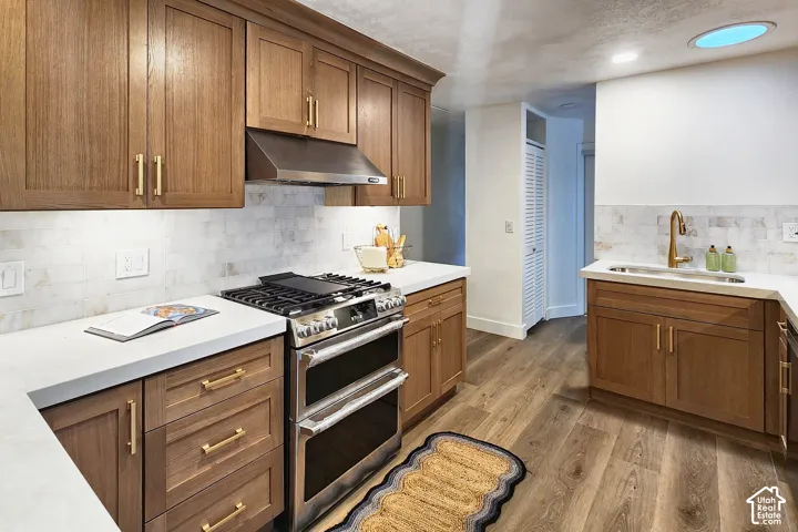 Kitchen featuring wood cabinetry with gold-tone hardware, white stone-finish countertops, subway tile backsplash, stainless steel gas range with a matching vent hood, and wood-finish flooring