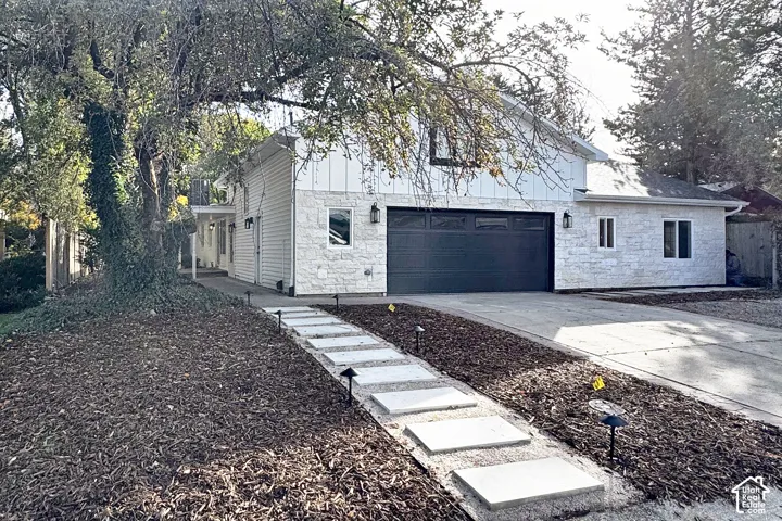 Exterior featuring white horizontal siding and stone masonry, a dark-toned multi-car garage door, integrated wall-mounted exterior lighting, and a concrete paver walkway