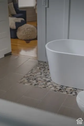 Freestanding white soaking tub set on a decorative river stone tile inlay, framed by large-format neutral floor tiling and board-and-batten wall detailing
