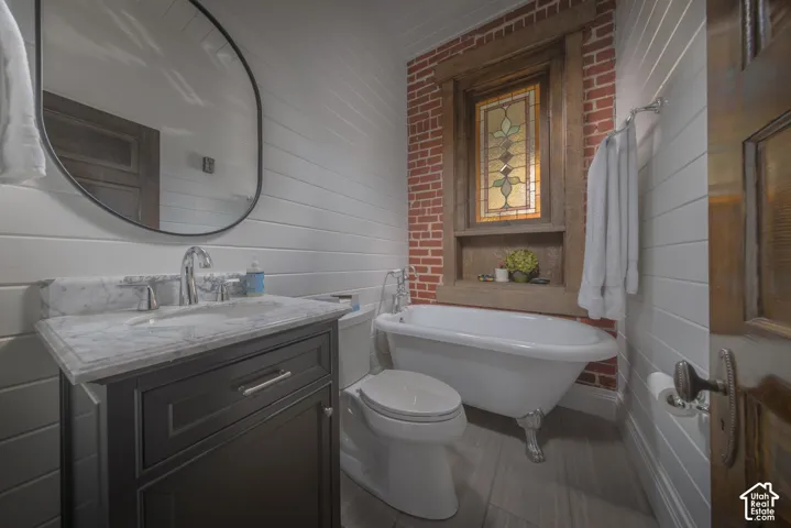 Clawfoot tub with polished hardware, exposed brick accent wall, stained-glass window insert, wood-plank wall paneling, and stone-top vanity