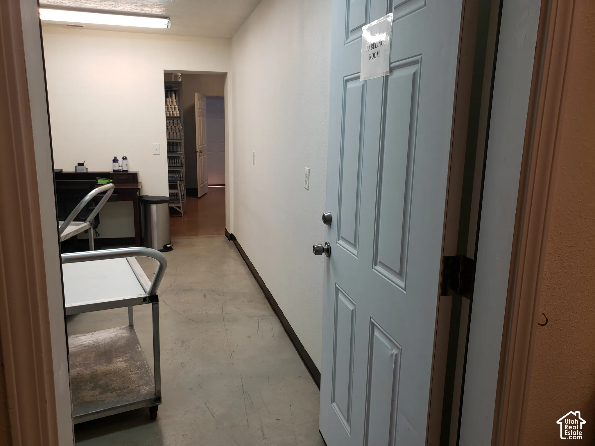 Interior corridor featuring smooth-finish concrete flooring, neutral painted walls, six-panel solid core door, and integrated overhead fluorescent lighting