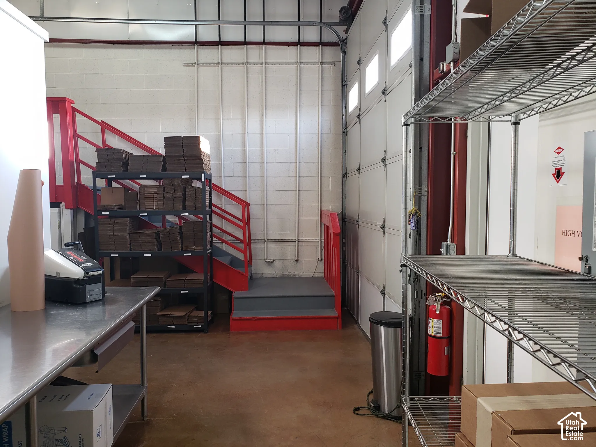 Industrial workspace featuring a high-clearance sectional roll-up door, exposed concrete flooring, white masonry walls, and a painted steel staircase with integrated railings