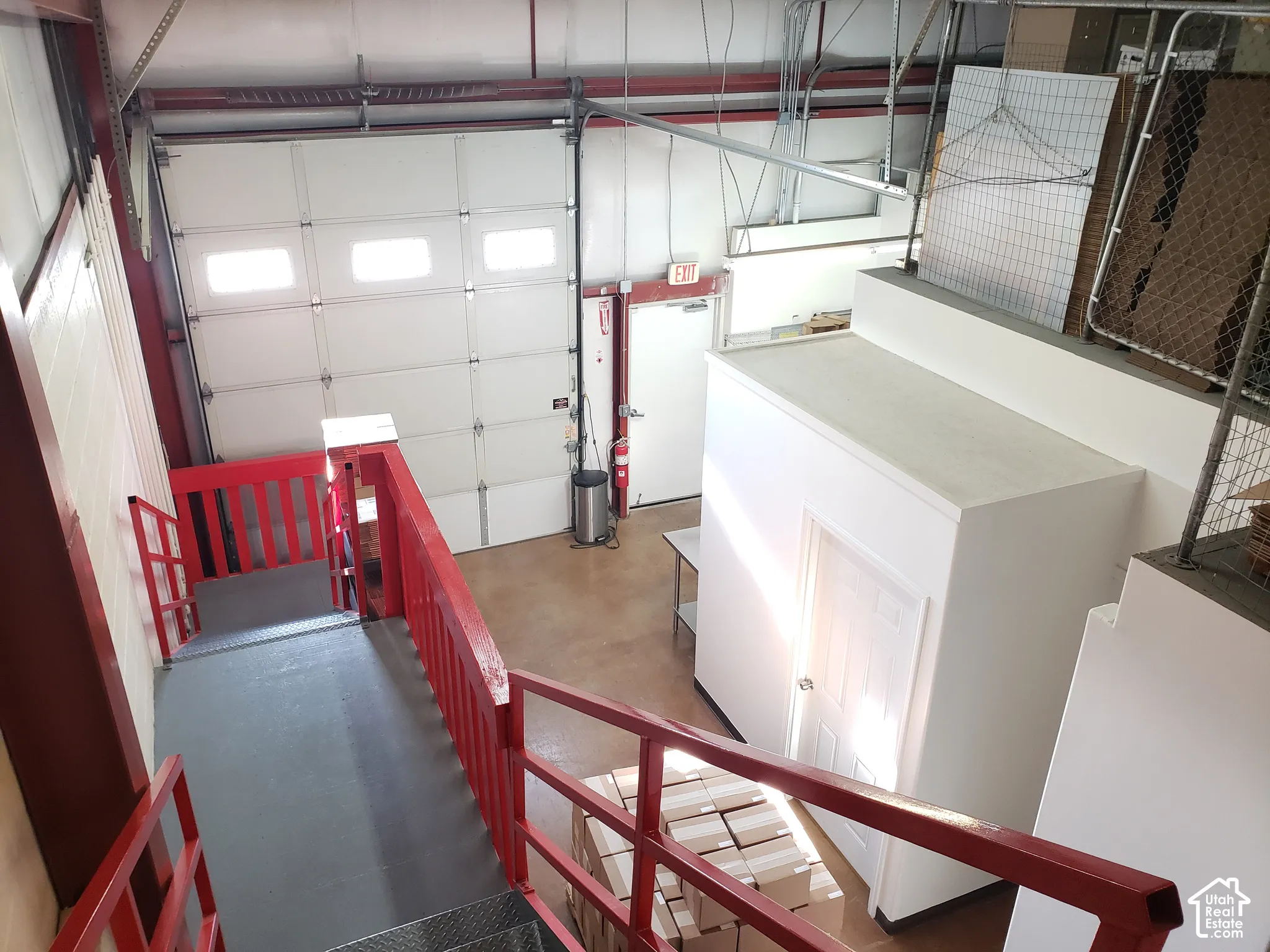 Industrial-style space featuring a high-clearance overhead garage door, concrete flooring, enclosed storage structure, and red-painted metal staircase railings