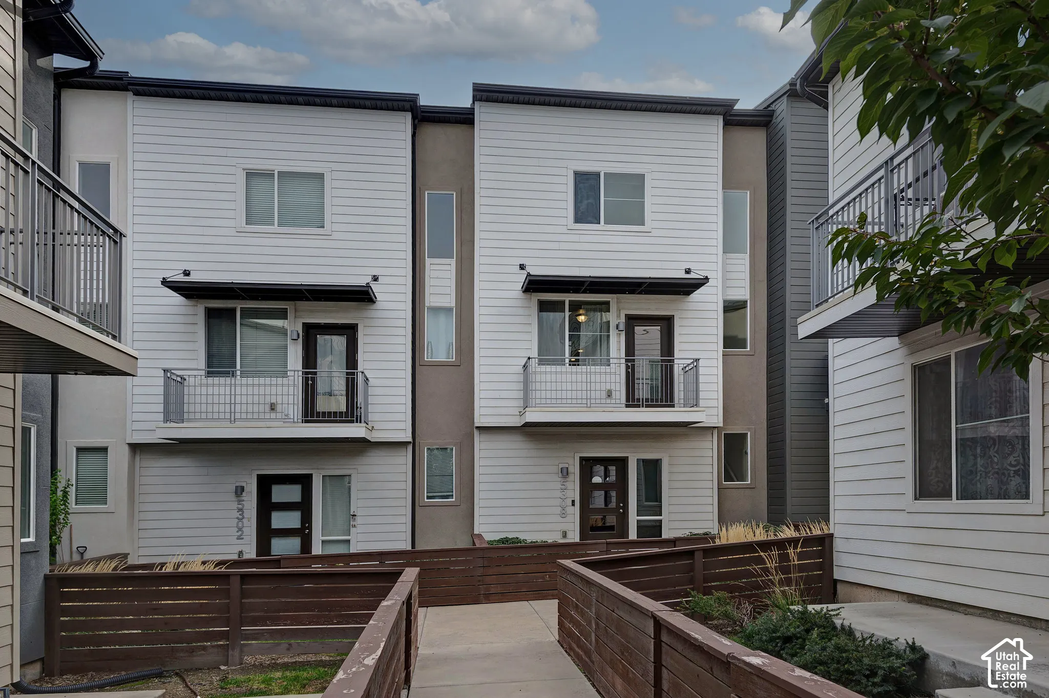 Multi-level exterior featuring white horizontal siding, contrasting dark-toned stucco accents, second-story balconies with metal railings, and wood-slat fencing