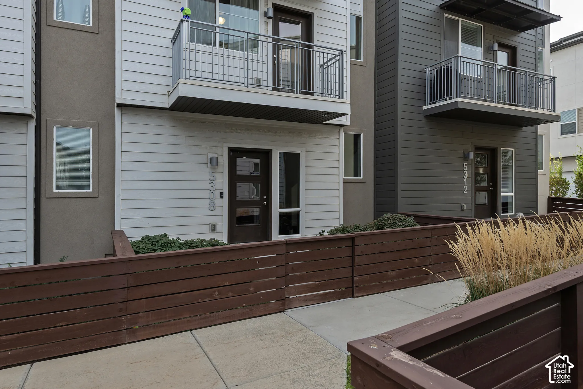 Modern exterior featuring horizontal lap siding, second-story balconies with metal railings, horizontal wood-slat fencing, and concrete hardscaping