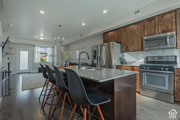 Kitchen featuring stained wood cabinetry, stone countertops, stainless steel appliances, a central island with a high-arc faucet, and wood-finish flooring