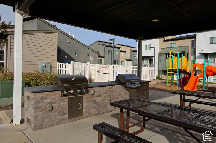 Covered outdoor kitchen featuring a stone-veneer island, dual stainless steel grills, and metal-frame tables on a concrete patio
