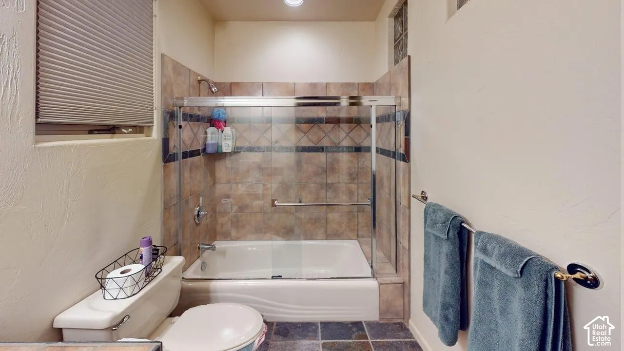 Bathroom featuring a tub-shower combination with tile surround and metal-framed glass sliding doors, stone-finish tile flooring, a horizontal window with a pleated shade, and built-in metallic hardware