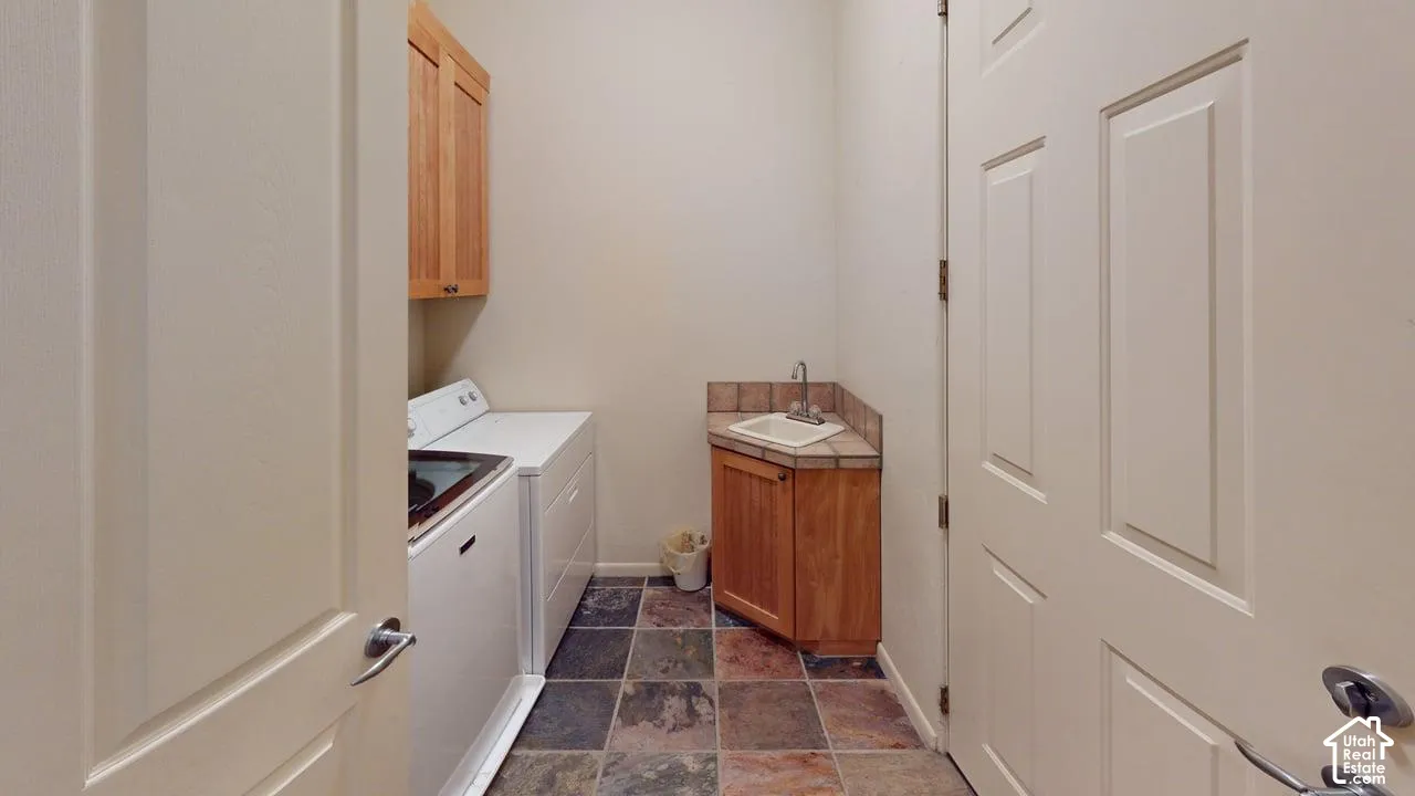 Laundry area featuring multi-tonal tile flooring, natural wood cabinetry, a corner utility sink with tile backsplash, and overhead storage cabinets