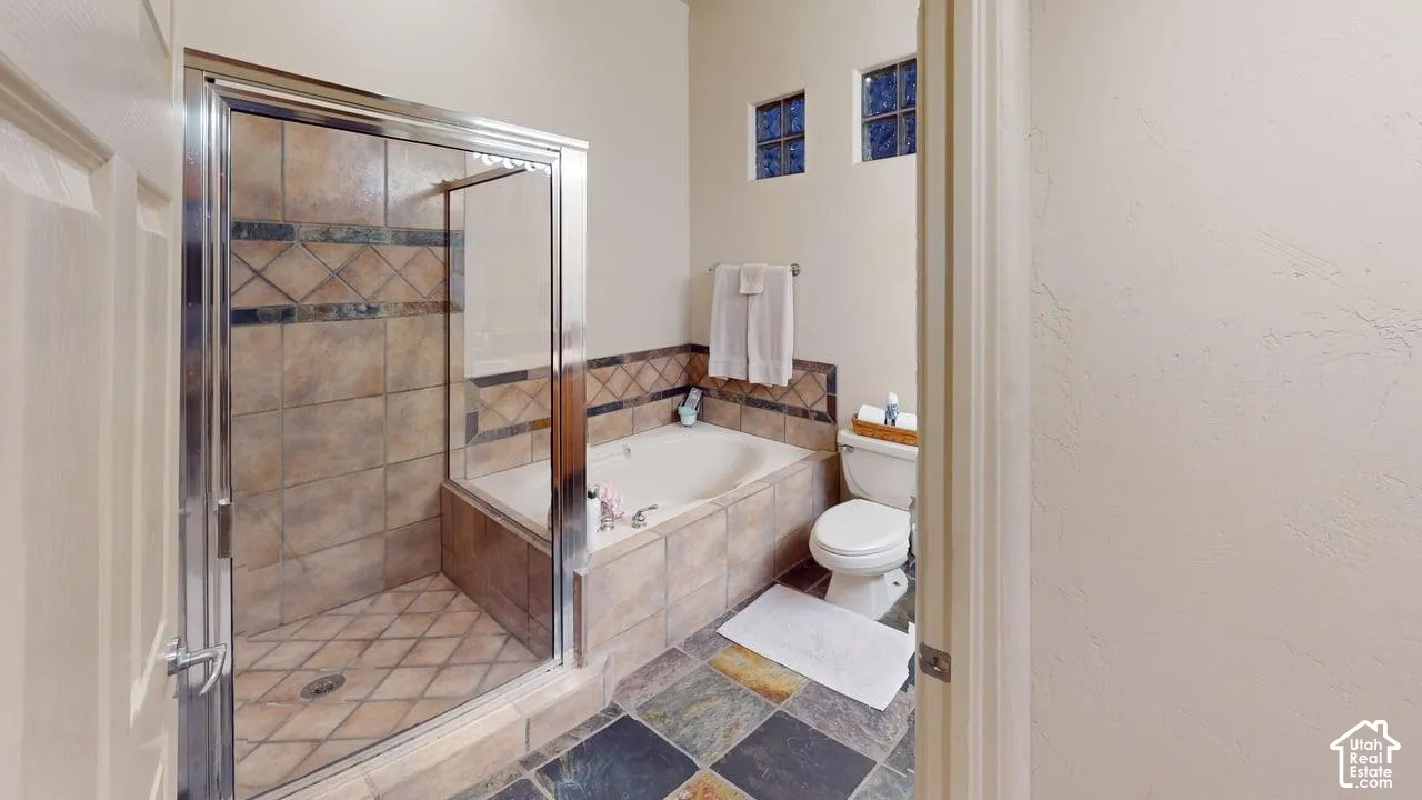 Bathroom featuring a walk-in shower with glass enclosure, tiled soaking tub, multi-colored stone flooring, and two high-set accent windows