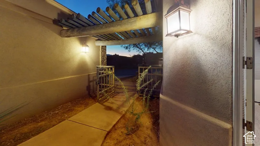Exterior entryway featuring stucco walls, a wood-beam pergola, concrete walkway, and integrated wall-mounted lanterns