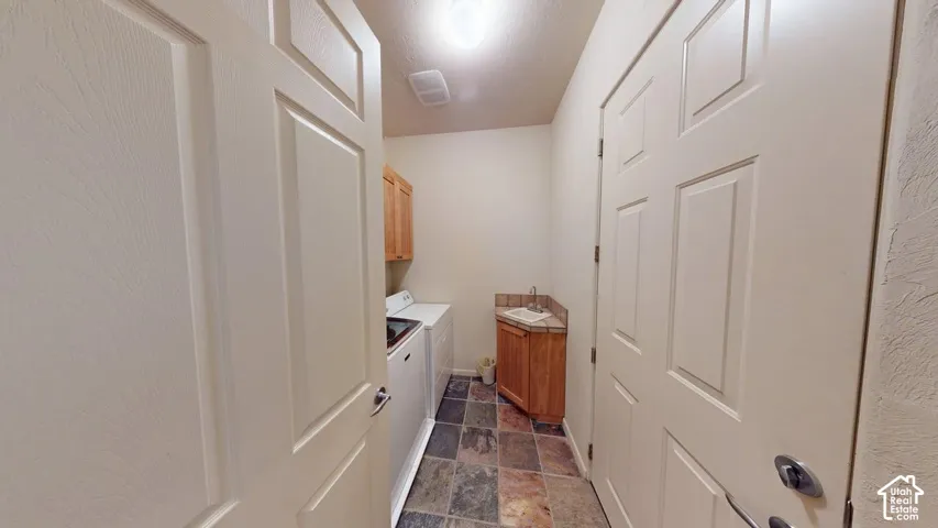 Laundry area featuring multi-tonal stone flooring, wooden cabinetry, and a corner sink with a tile backsplash