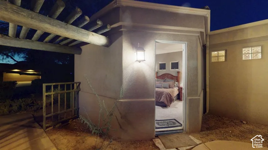 Stucco exterior featuring a timber beam pergola, metal entry gate, wall-mounted lantern-style lighting, and glass block accent windows