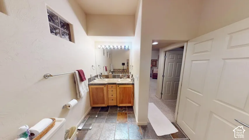 Dual-sink vanity with light wood cabinetry, stone-tiled countertop, wall-to-wall mirror, and slate flooring