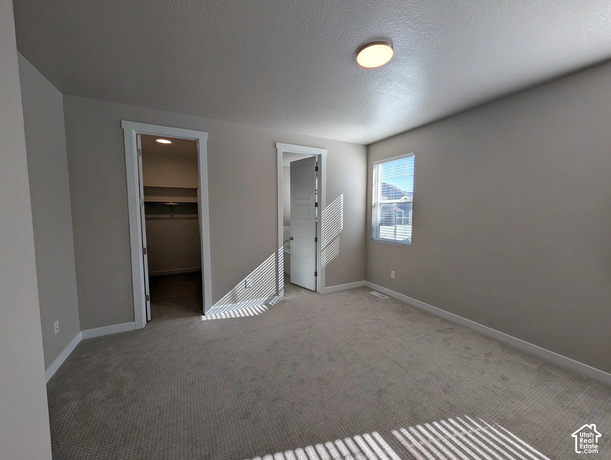 Neutral-toned carpeted room featuring a ceiling-mounted light fixture, one window, and two separate interior doorways leading to closet and room spaces
