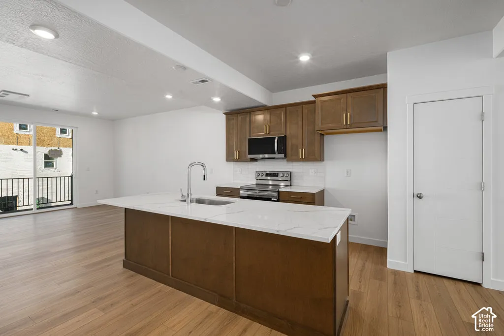 Kitchen featuring wood-finish cabinetry, stone-finish countertops, an island with an integrated sink, stainless steel appliances, and wood-finish flooring
