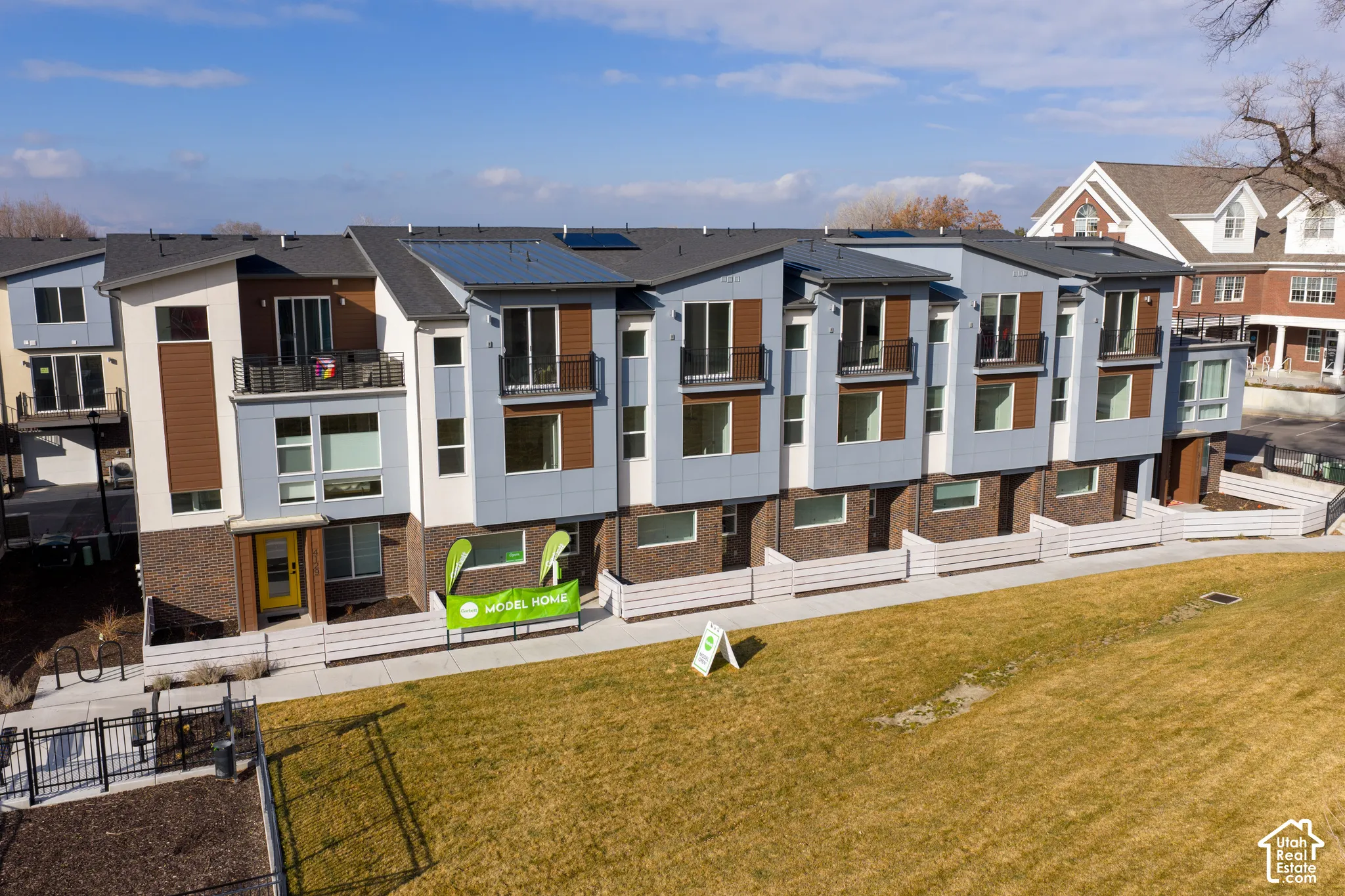 Multi-level townhome exterior featuring brick and panel siding, roof-mounted solar panels, private balconies with metal railings, and concrete walkways