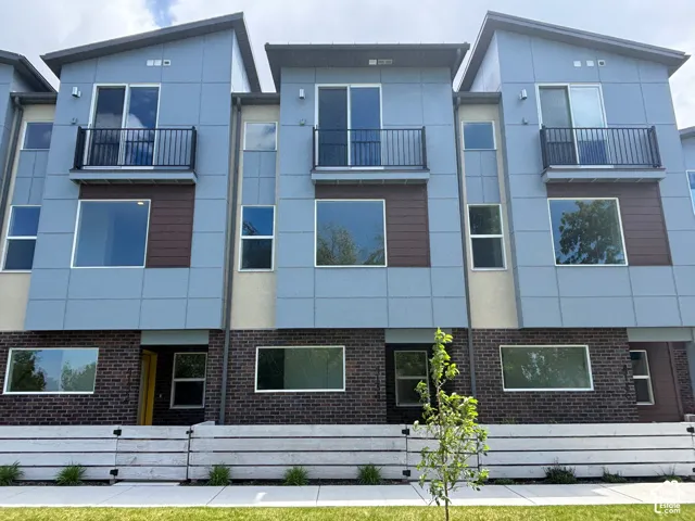 Multi-story modern townhouse exterior featuring composite panel siding, brick veneer cladding, metal railing balconies, and horizontal slat wood-finish fencing